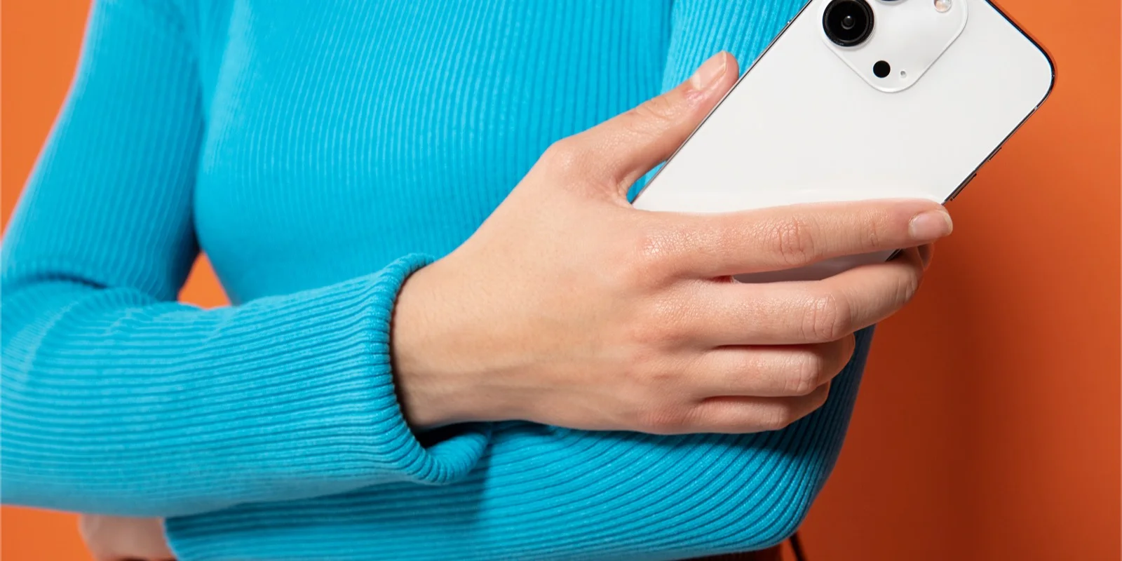 Una mano sostiene un teléfono blanco sobre un fondo naranja intenso, con el brazo en un suéter cian