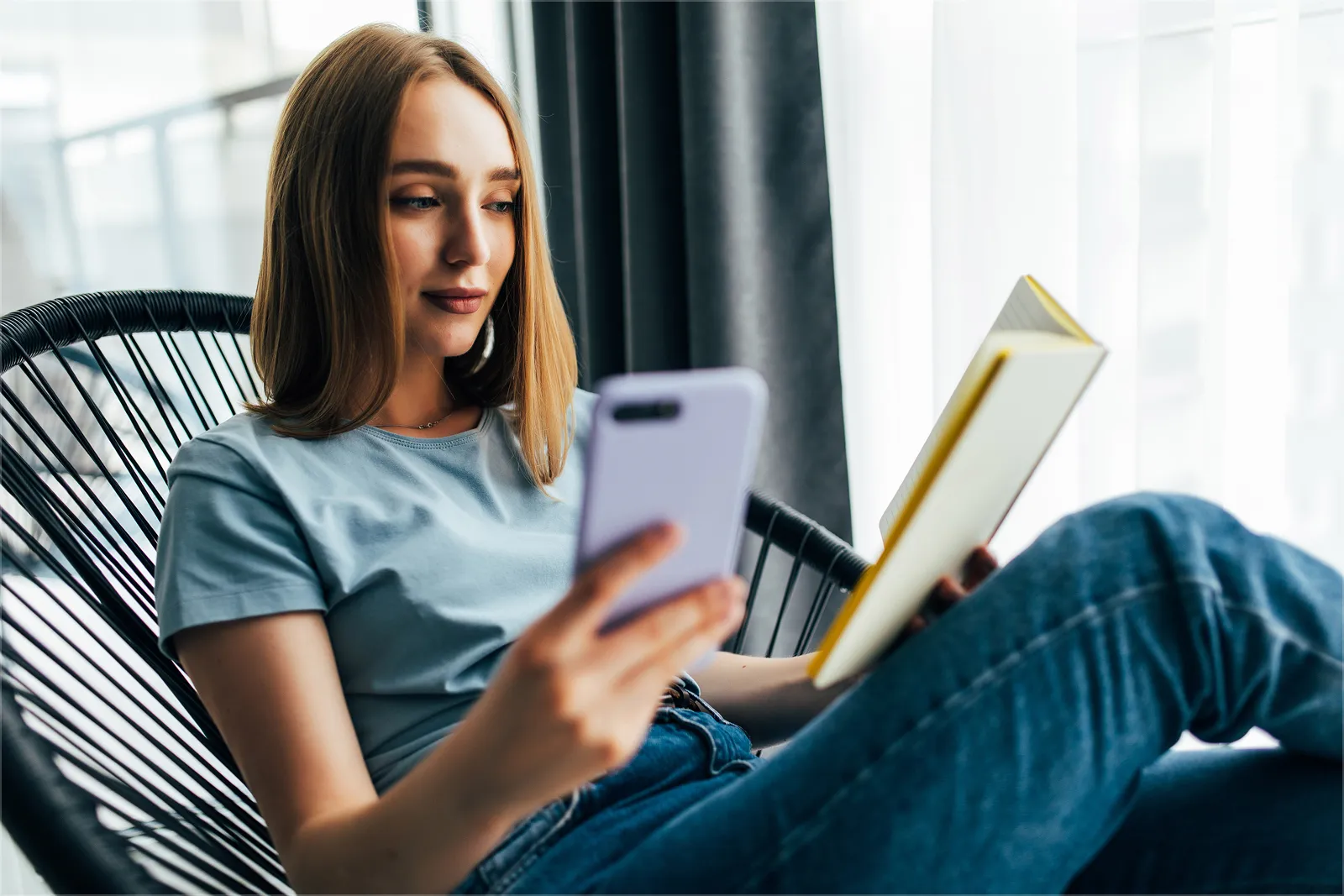 Una mujer sentada en un sillón de mimbre negro junto a una ventana, con un teléfono en una mano y un libro abierto en la otra